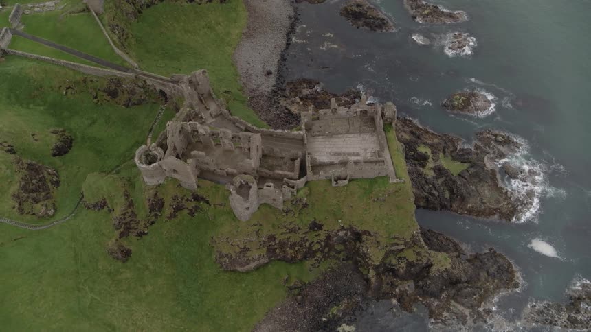The ruins of a stone castle are perched atop a rocky cliff over the ocean on a grass covered coastline