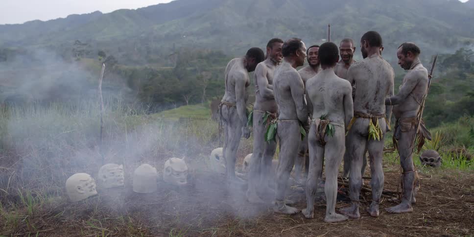 Several african men stand in a circle around a smouldering fire with clay masks resting on the ground nearby