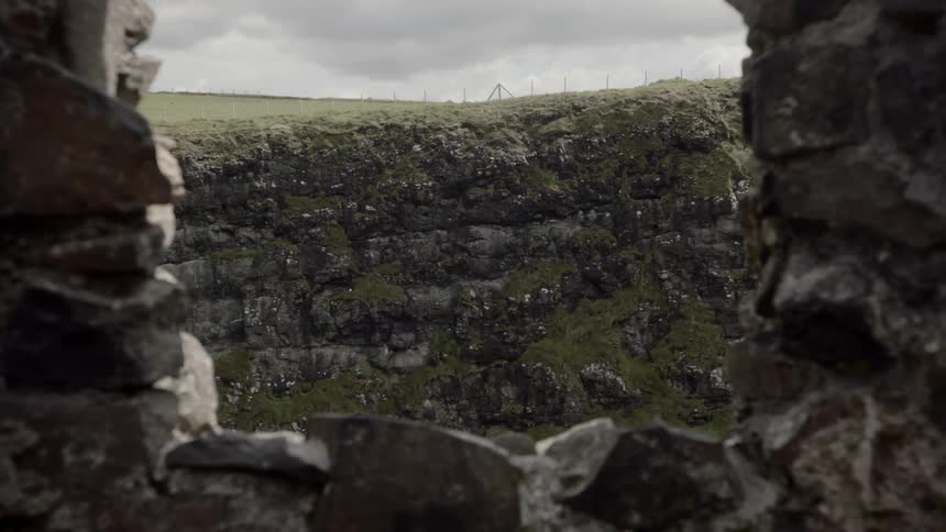 Through a window cutout of a stone wall a very stony cliff face can be seen with smooth grassy fields atop it
