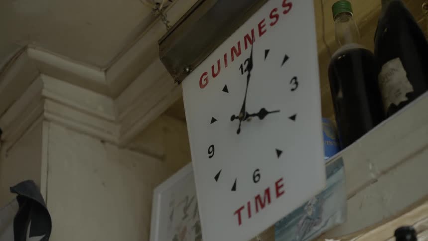 An old fashioned clock hangs from the ceiling by two small chains inside of an older looking room and it displays the phrase guinness time on it