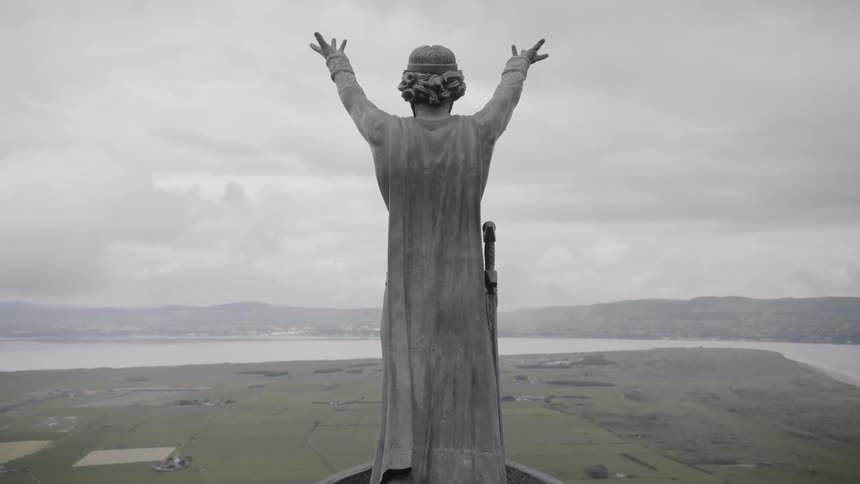 A statue with outstretched arms a sword at their side and a cape stands high above a flat valley at the edge of a body of water