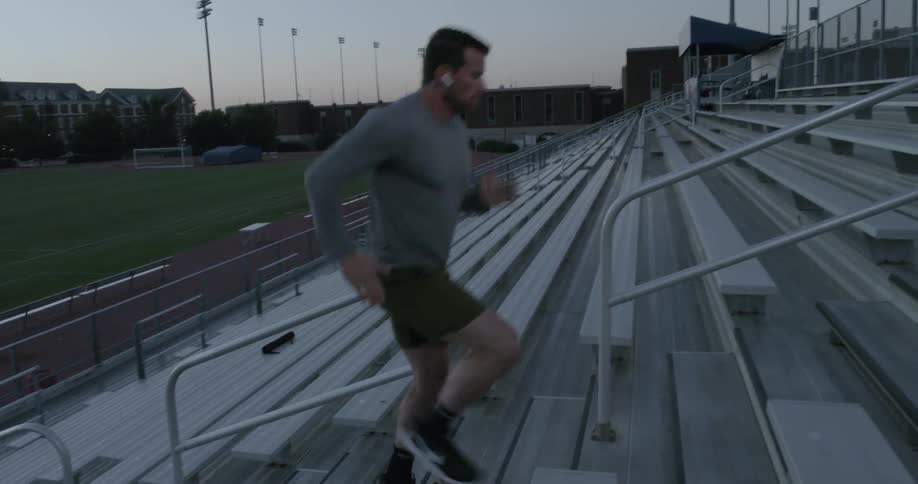 A white man in a gray sweatshirt and green shorts runs up bleachers