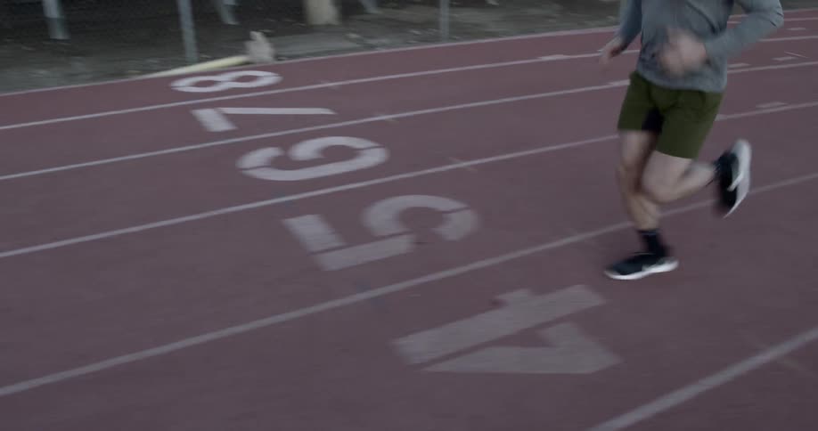A white man in green shorts and a gray sweatshirt jogs along a stadium racing track
