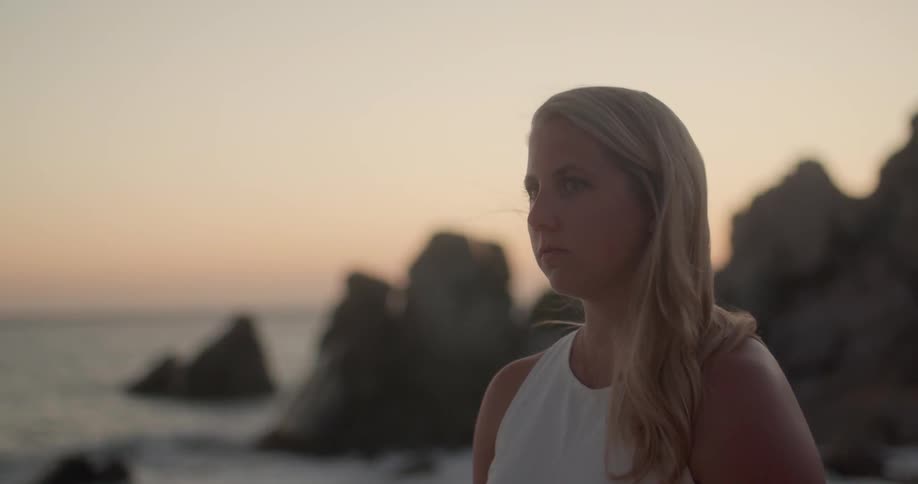 A young white woman standing on a shoreline at sunset