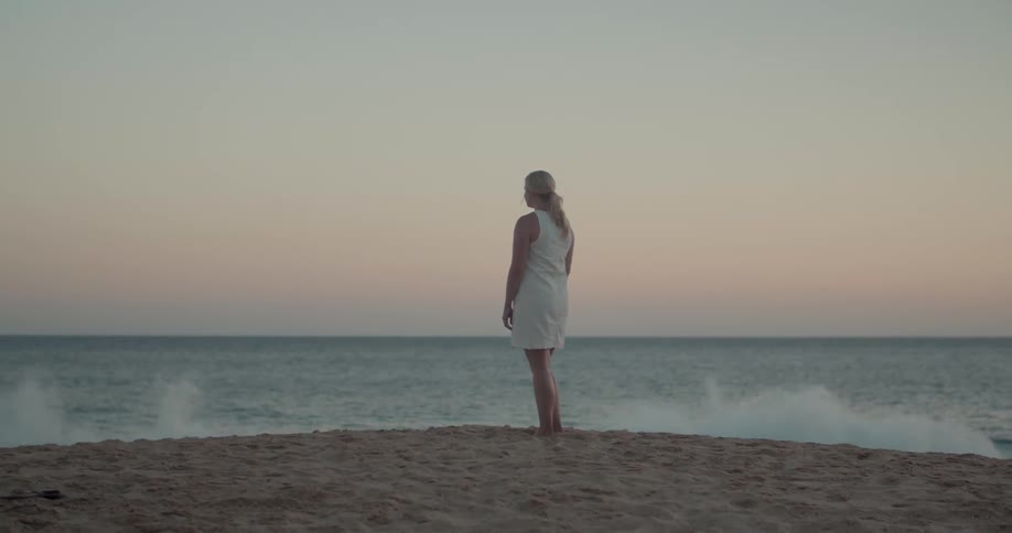 Woman stands on beach looking out to sea at sunset