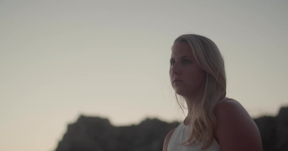 A blond female looks contemplative and then turns her gaze downward with cliffs in the distance behind her