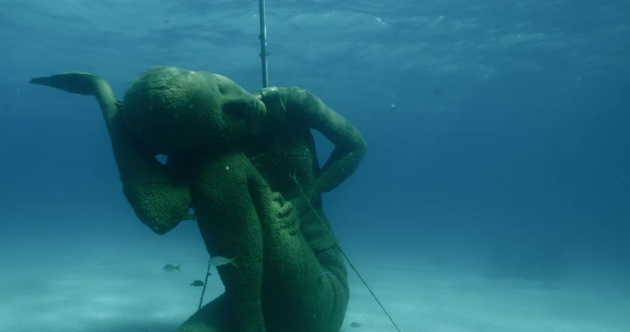 A mossy statue of a person sits on a seabed underwater