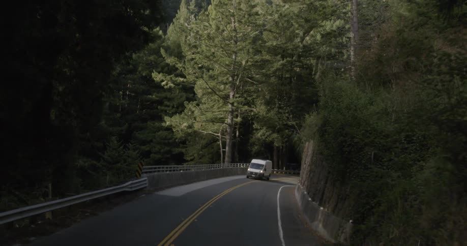 A truck travels on a narrow rural road lined with trees and a slope