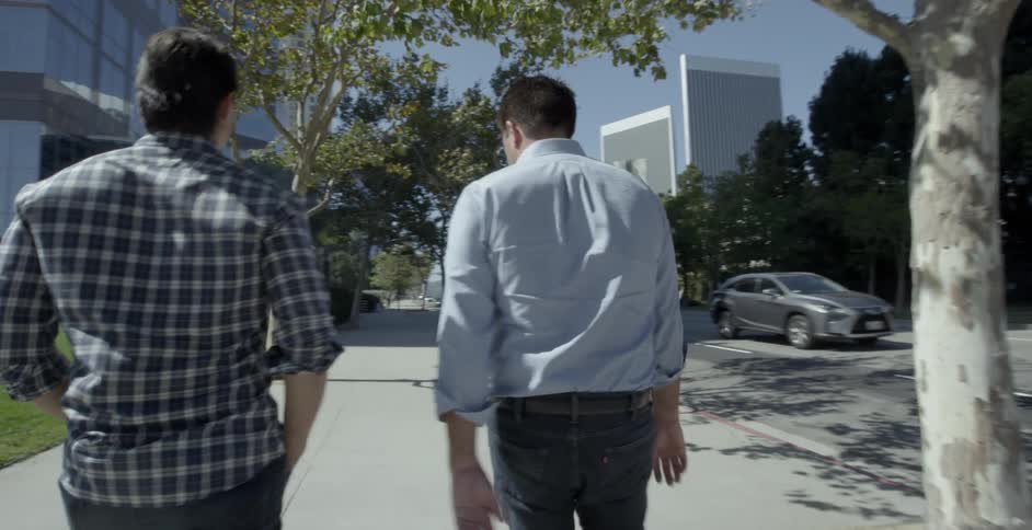 Two white males in button down shirts and jeans are walking down a treelined sidewalk of a downtown area on a sunny day
