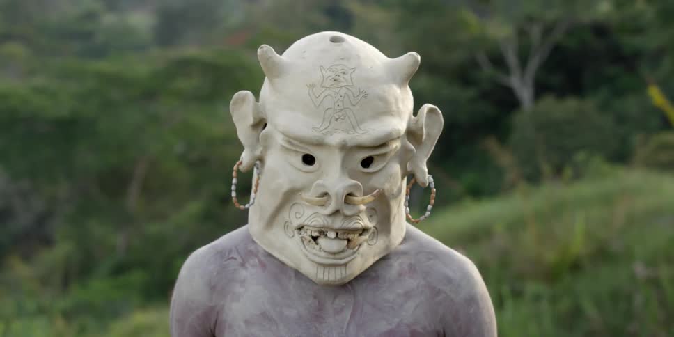 An african man covered in a thin layer of clay wears a large clay mask over his head