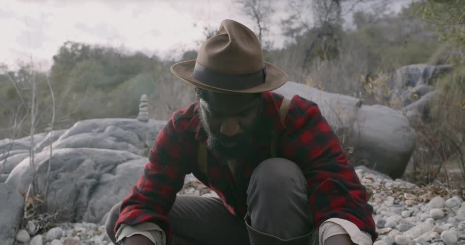 A man in a red plaid shirt and black boots pans for gold next to a river