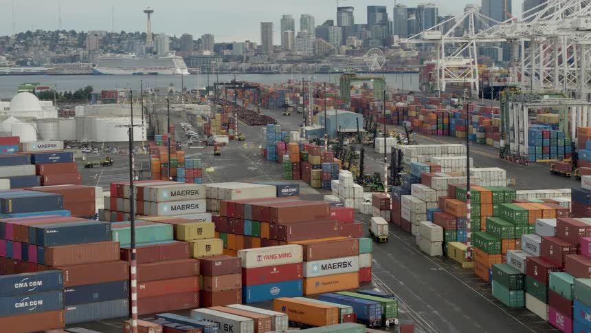 A truck takes a turn in a shipyard full of stacked shipping containers and cranes on the waterfront