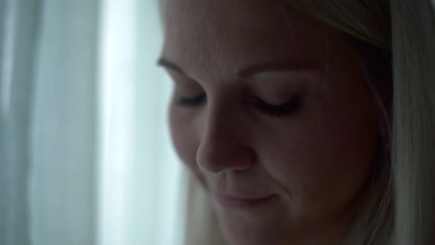 A blonde female is looking down and smiling while standing near a window