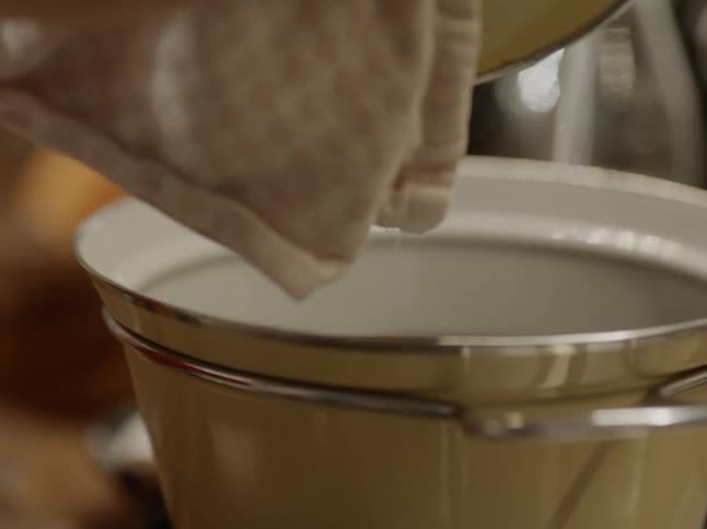 A person uses a towel to lift a lid from a hot pan on a stove