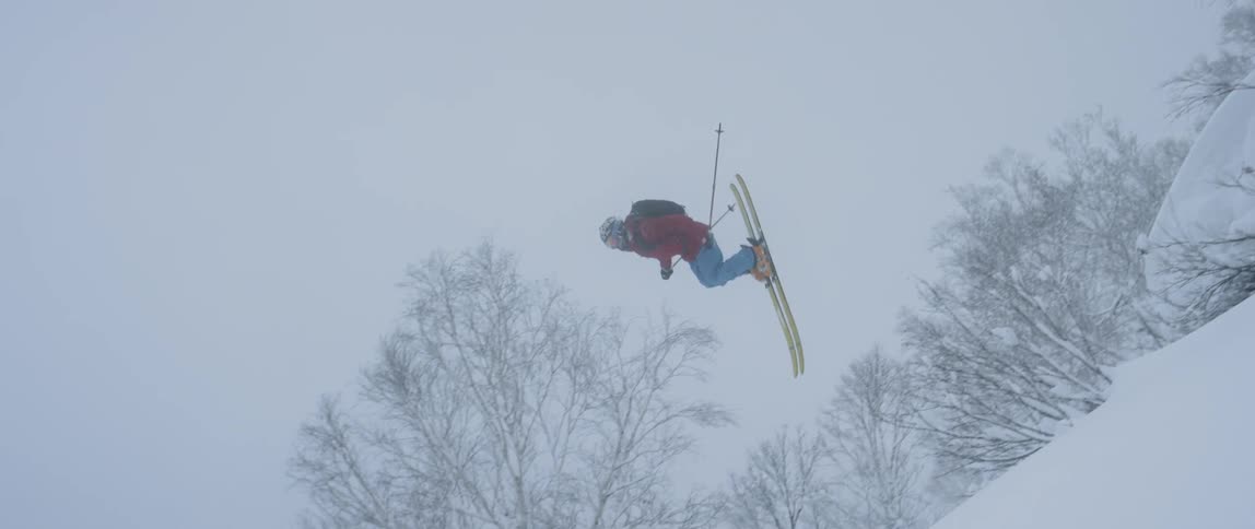 A skier doing a back flip on a ski slope surrounded by snow and trees