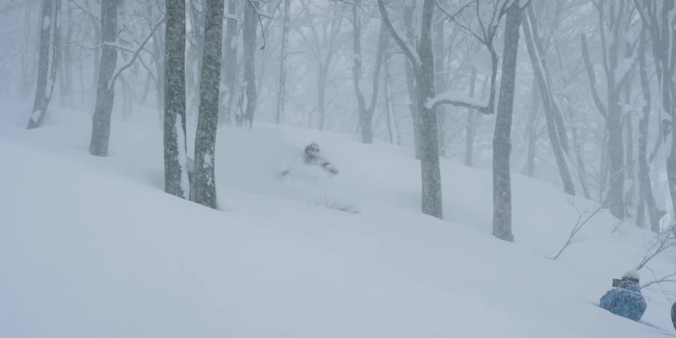 A skier skis down a slope through fresh powder while people film him