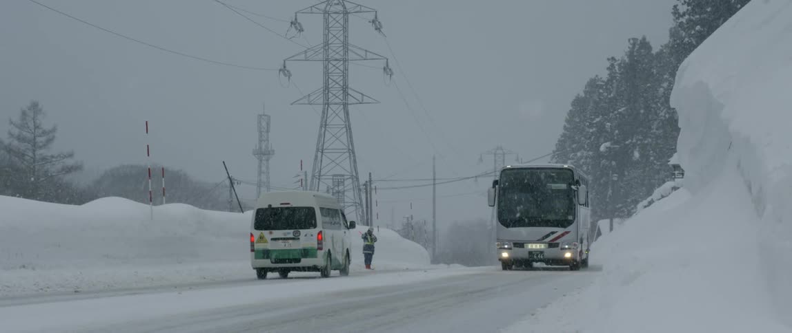 A person directs traffic in the snow on the side of a road as buses pass