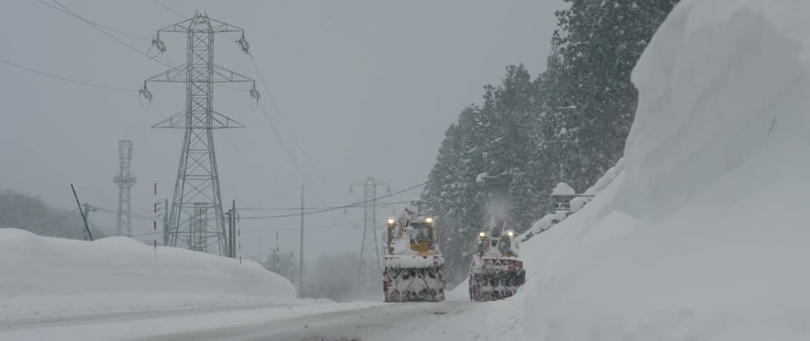 Two large construction vehicles move down a snow covered road