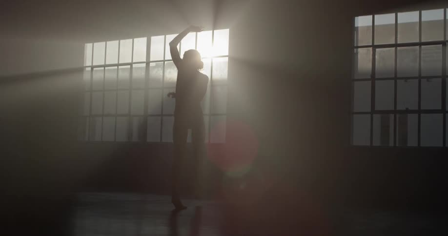 In a dimly lit studio a dancer is seen dancing through the sun casted rays peaking through the windows projecting a flare like shadow
