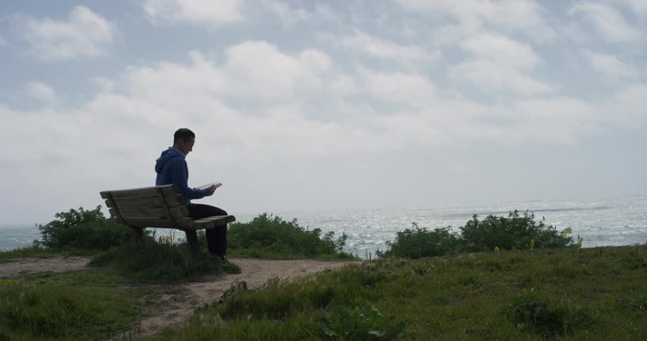 Man sits on bench overlooking the ocean while reading a book