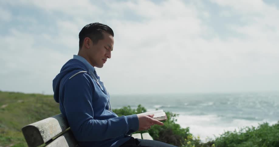 A man reads a book while sitting on a park bench along the coast