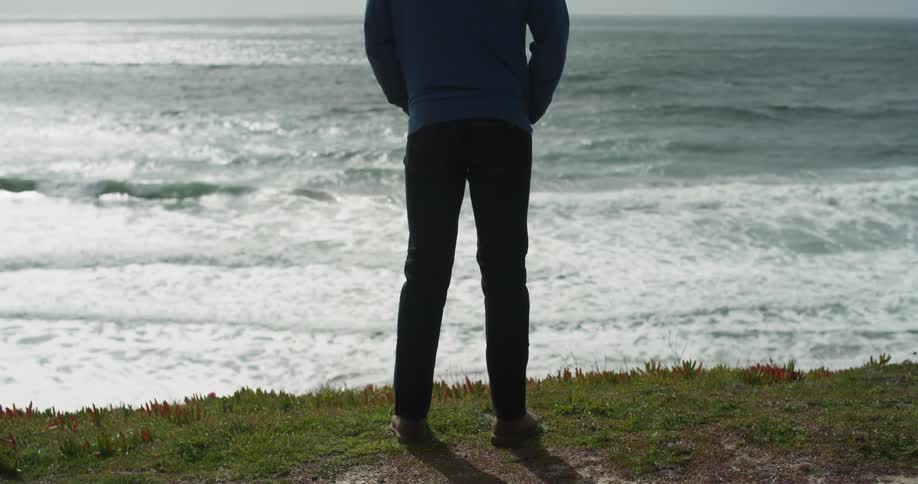 A man with a blue hoodie standing watching the ocean