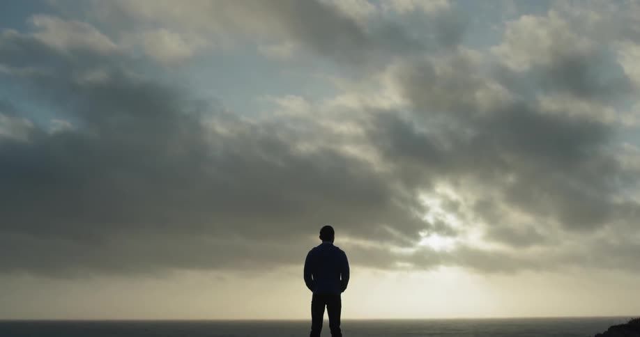 Under the cloudy sky a young man stands observing the sunset