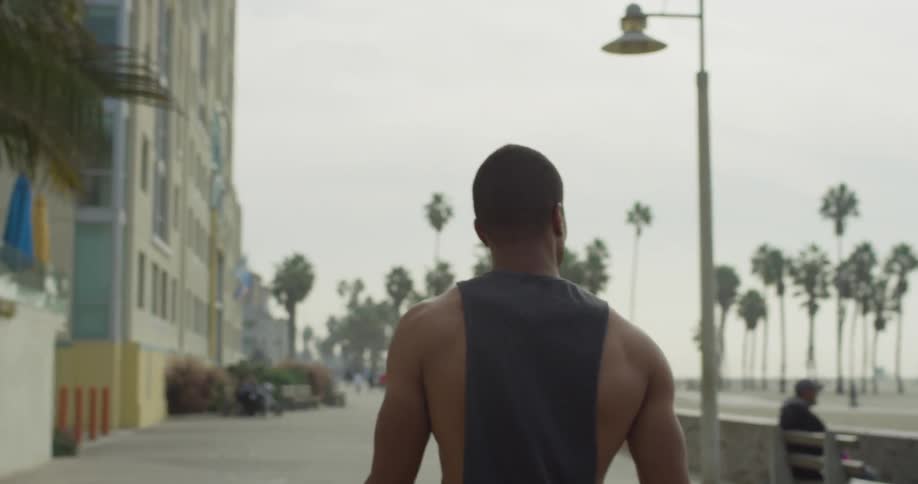 A muscular man in a tank top walks on a boardwalk on a beach