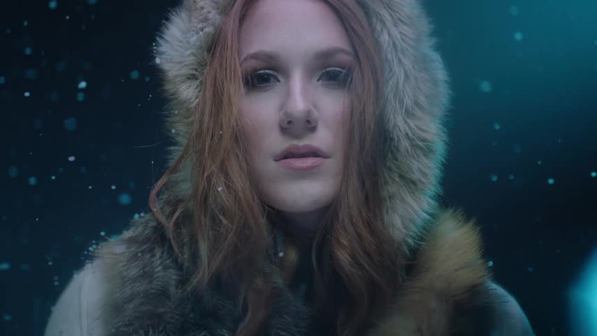 A woman wearing a fur lined hood stares into the camera while snow falls