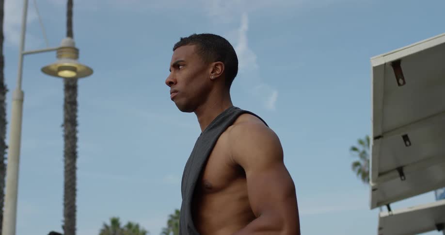 A muscled man stands on a boardwalk under a street light and palm trees
