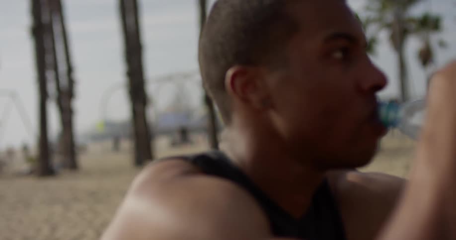 A black man sits on the beach and drinks water while people frolic in the background