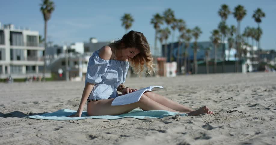 Woman sits on the towel laid on the sand beach and leafs through a book on a sunny day