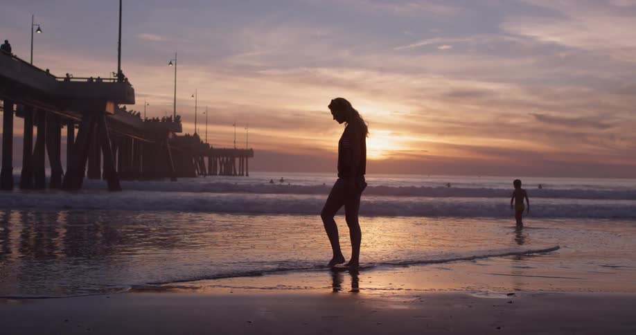 The silhouette of a woman can be seen against a setting sun a beautifully colorful sky as she walks on the beach and dips her toes into the shallow water of an small ocean wave while a child plays in the ocean waves beyond her