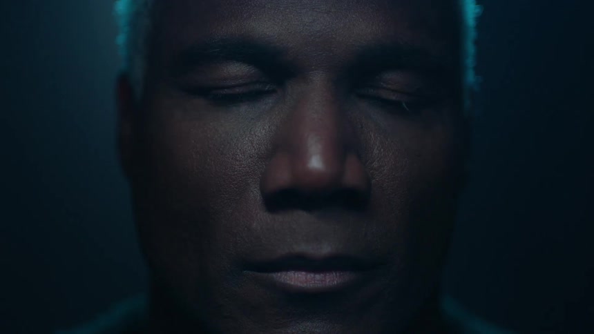 A black man with white hair blinks in a dark room