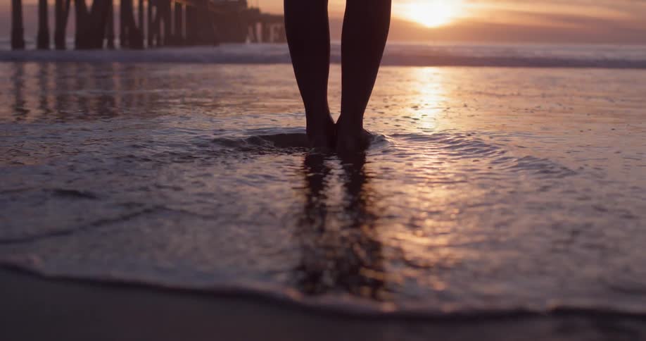 A woman stands on a sandy beach as the sun hangs low in the sky while the ocean tide washes over her feet