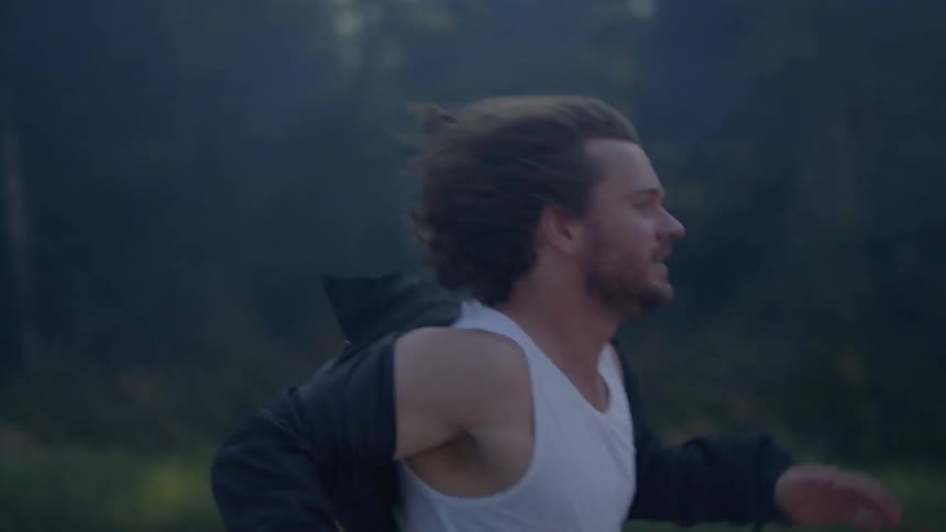 A young man runs by a wooded area with smoke in the air
