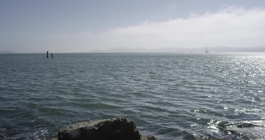 Ocean is gently rippling near rocks while a sail boat cruises by in the distance