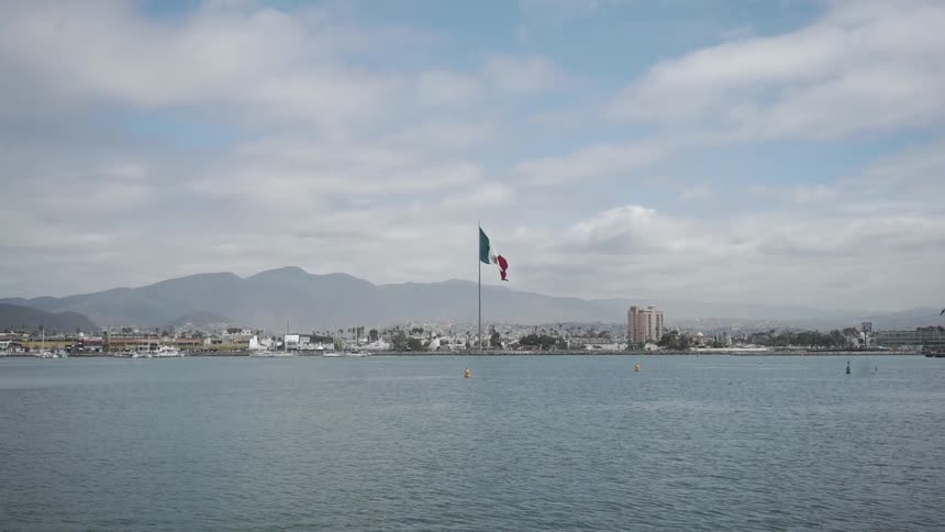 A mexican flag atop a flagpole at a harbor in a coastal town with mountains in the background in daylight