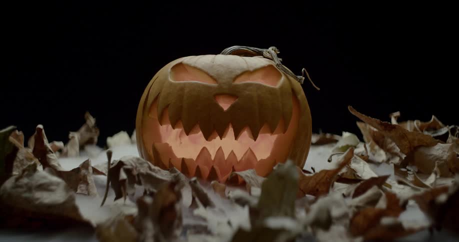 A glowing jack o lantern with a jagged smile sits on a bed of leaves with dry ice fog flowing around it