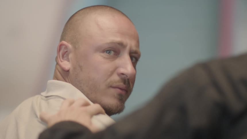 A guard handles a man out of a room as the apprehended man looks back with sorrowful eyes on what he leaves behind