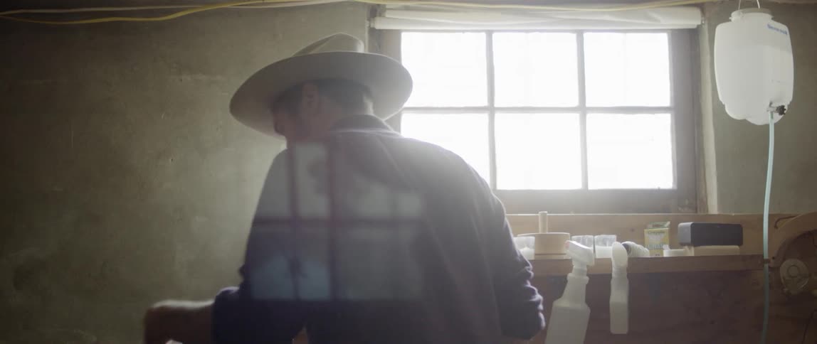 A man wearing and holding a hat is surrounded by bottles and containers of liquid