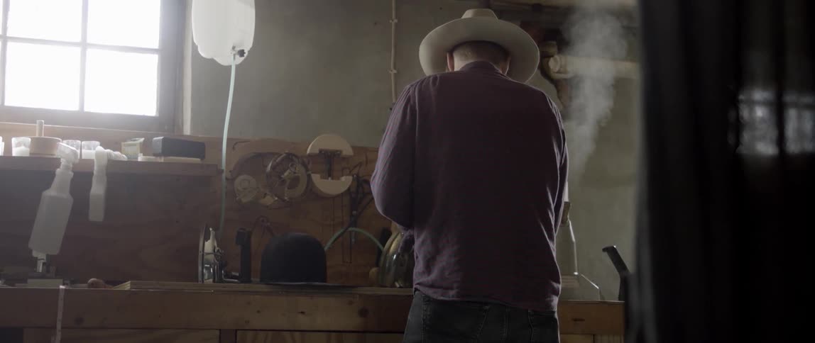 A white man wearing a hat works at wooden workbench