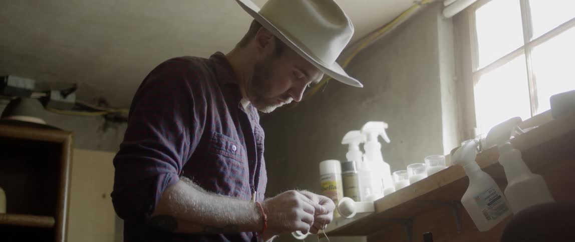 A milliner adapts a thread in his workshop next to shelves underneath a window on a sunny day