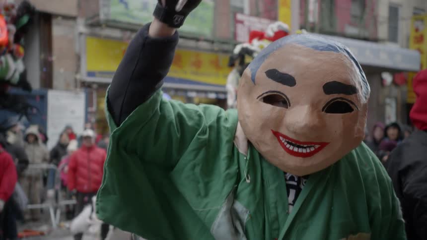 A person wearing a large fake costume head made out of paper turns and waves a fan amidst other performers wearing Chinese dragon costumes in a street parade