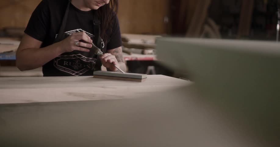 White woman with long brown hair and tattooed arm in black shirt is sitting down sharpening her chisel