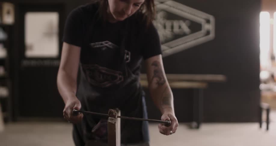 A woman uses a carving tool to remove the end of a wood post