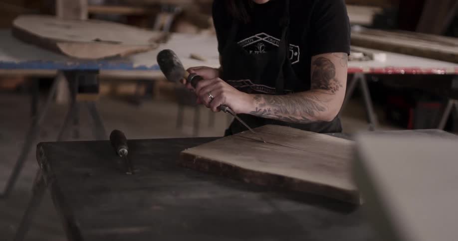 A woman with tattoos uses a chisel and hammer on a piece of wood in a workshop