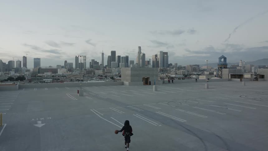 A young man walks while bouncing a basketball on an empty rooftop parking lot