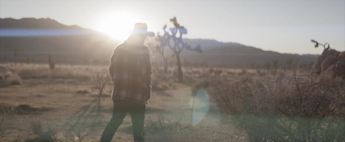 Man wearing wide hat walks on dry grass while the sun goes down behind the mountains