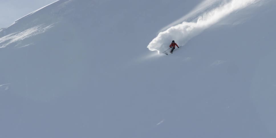 A skier skis down a steep slope through fresh powder on a sunny day